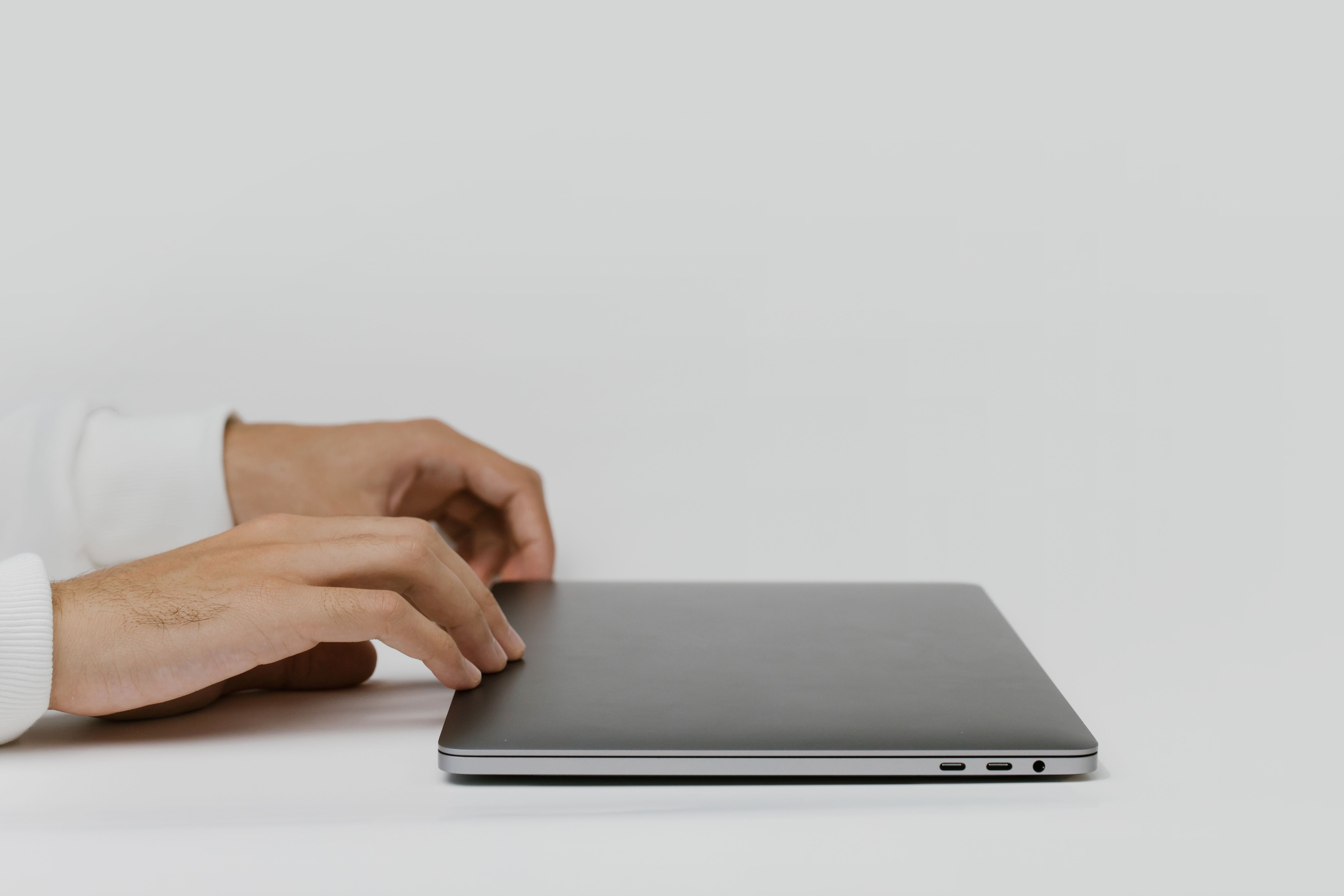 Hands about to open a closed silver laptop on a white desk with a plain background.