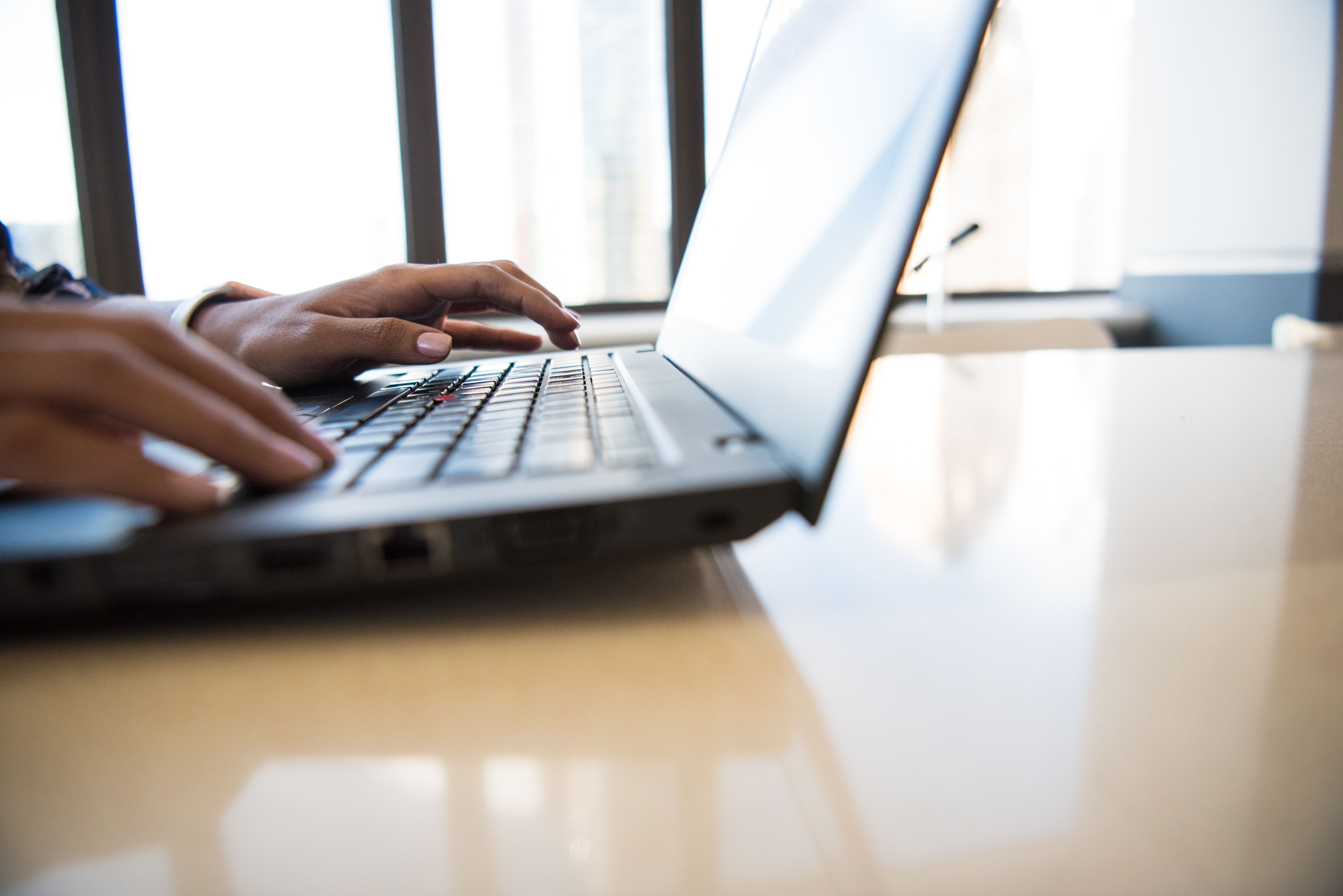Hands typing on a laptop keyboard beside a large window with city view.