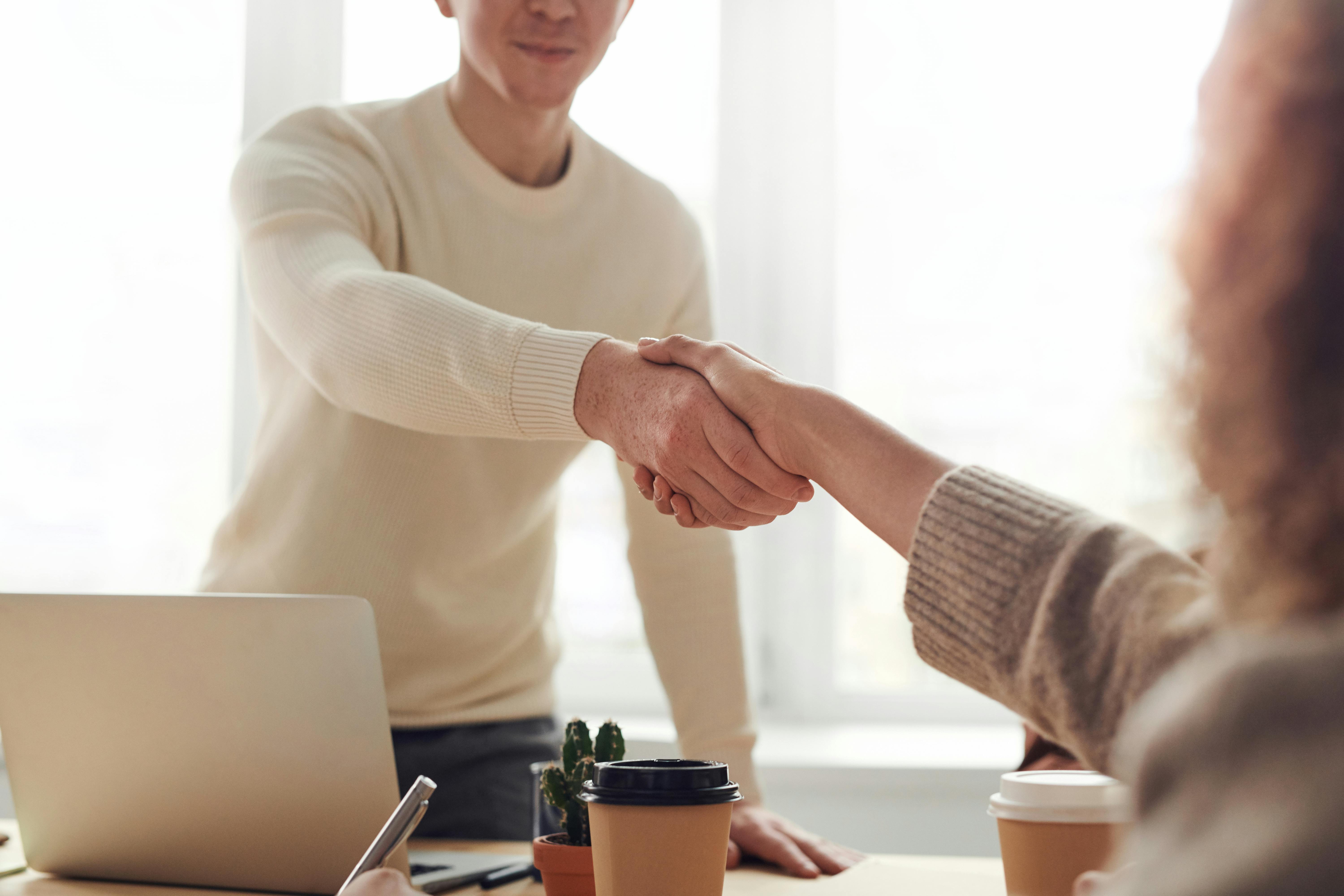 Two people shaking hands over a table with coffee cups and a laptop in a bright office.