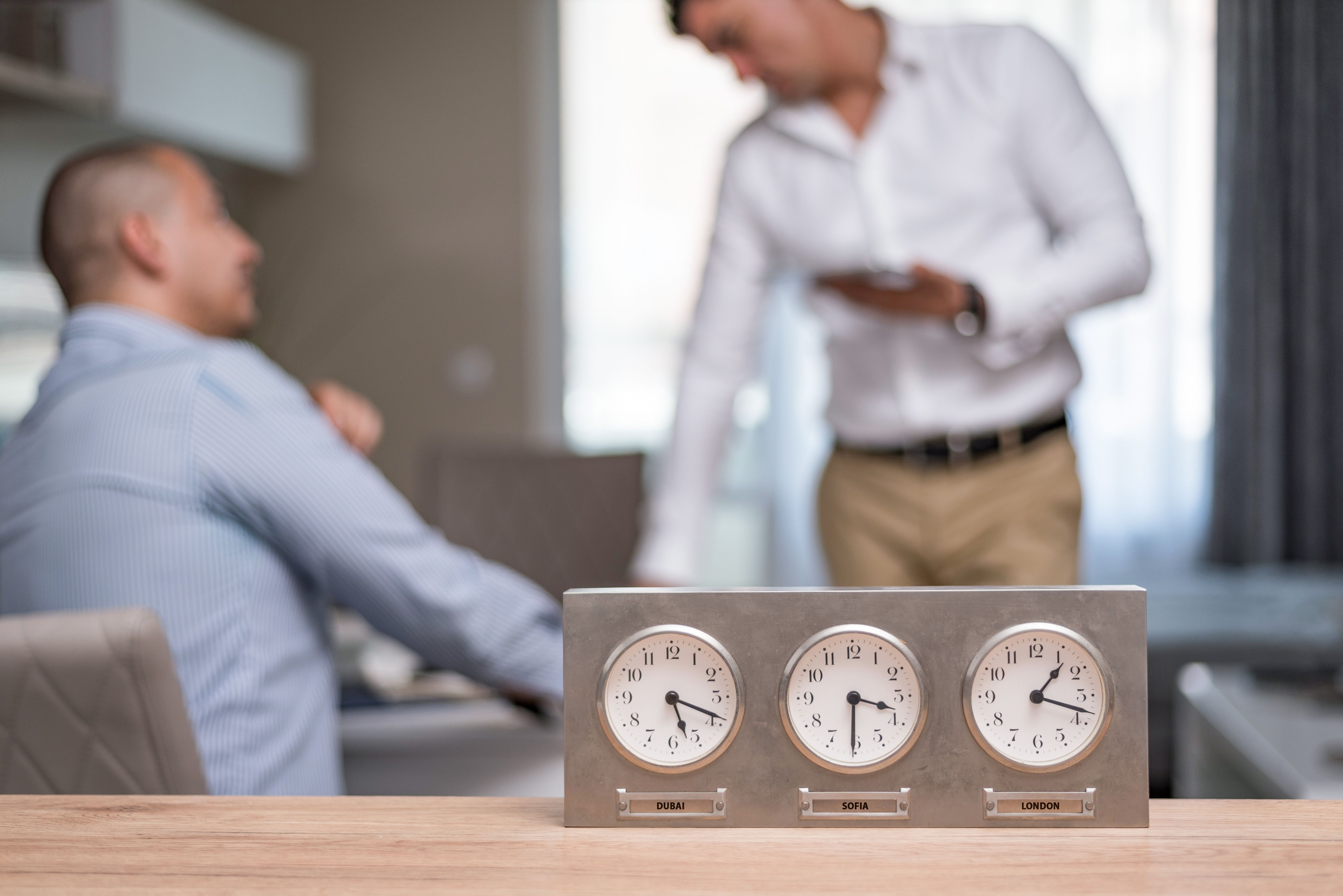 Three desk clocks showing times in Dubai, Sofia, and London with two blurred men in background.