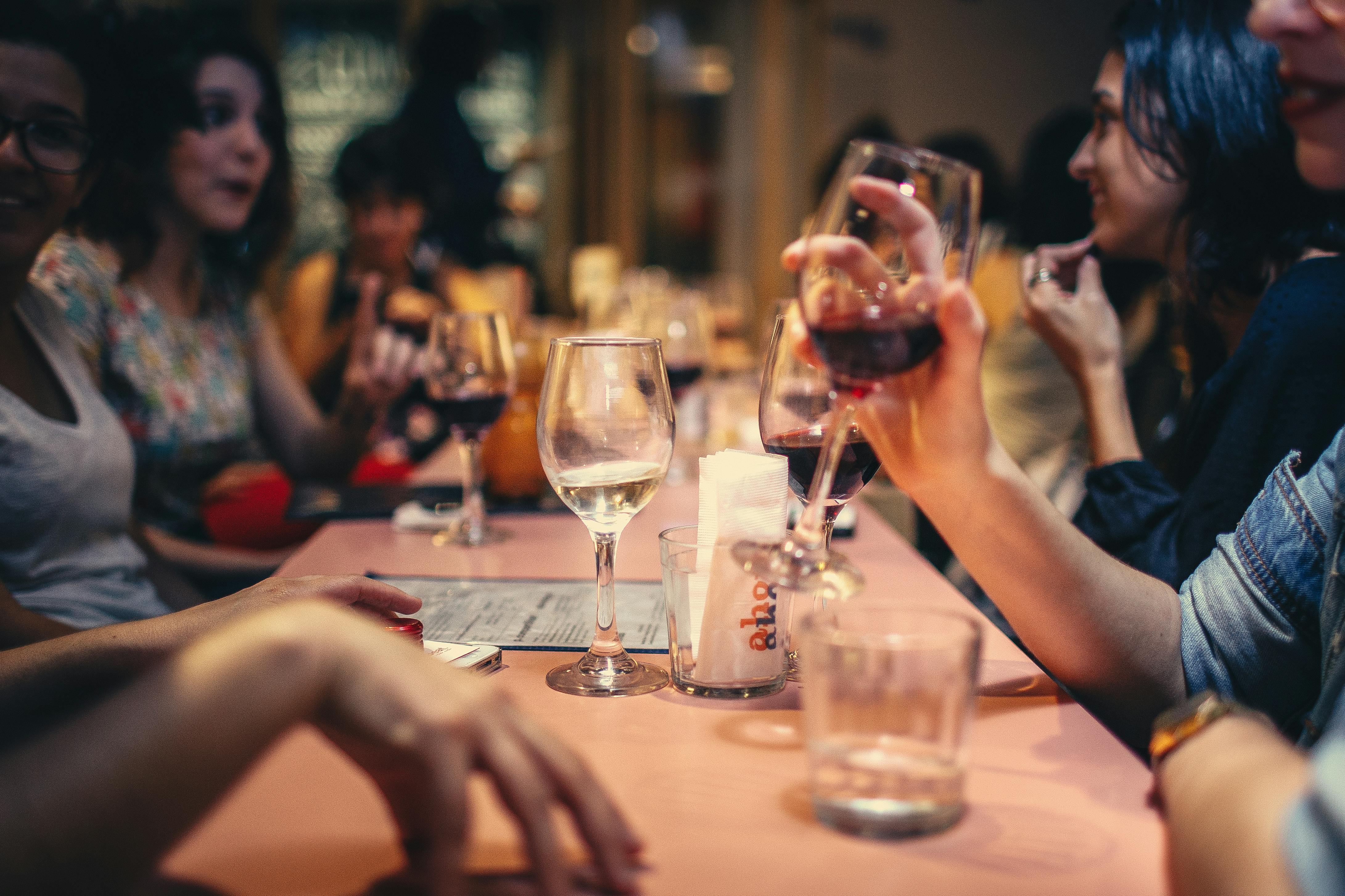 Group of people sitting around a table with wine glasses, drinking and chatting in a dim setting.