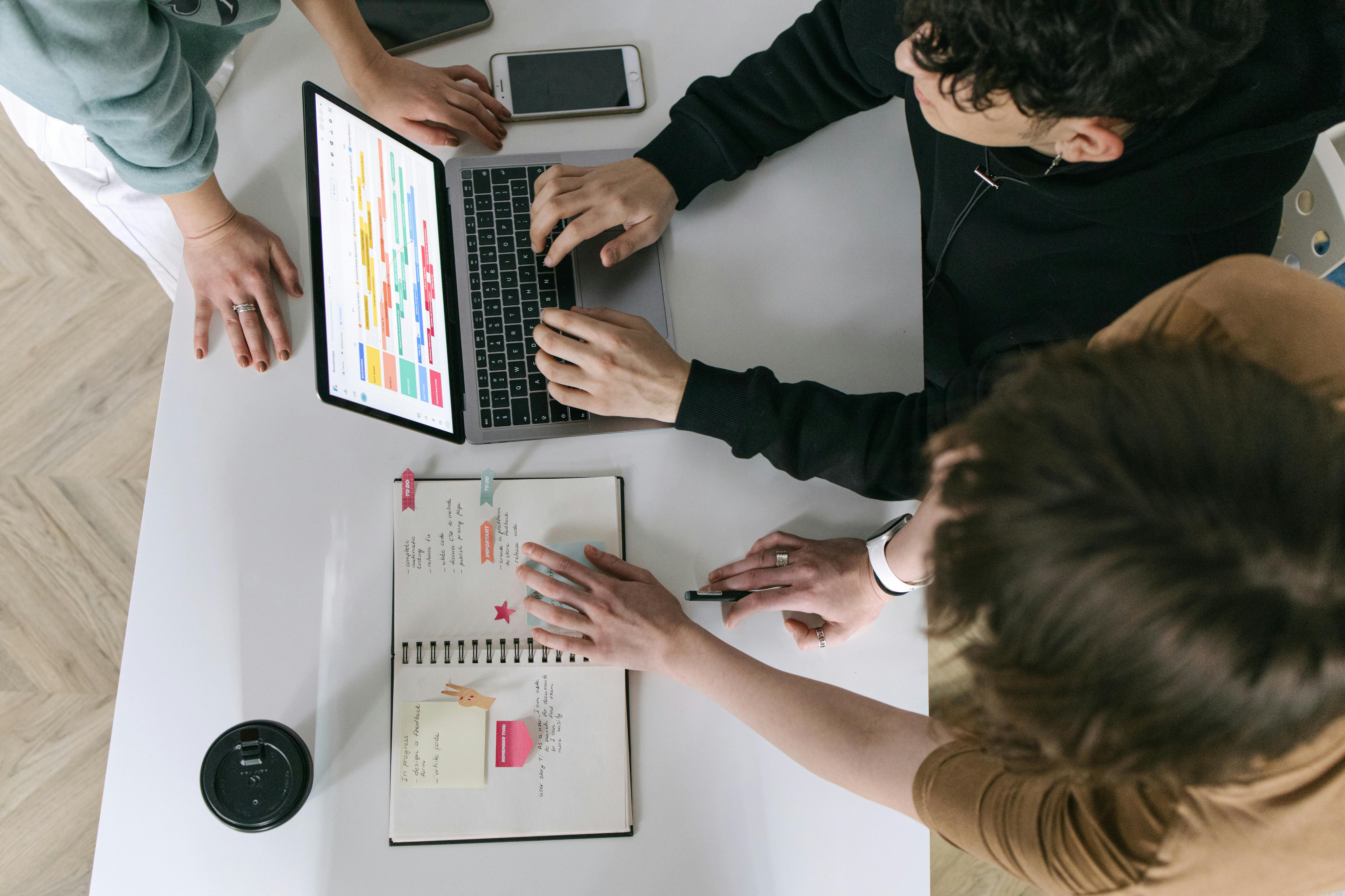 Three people collaborate at a table with a laptop showing a colorful chart and an open notebook.