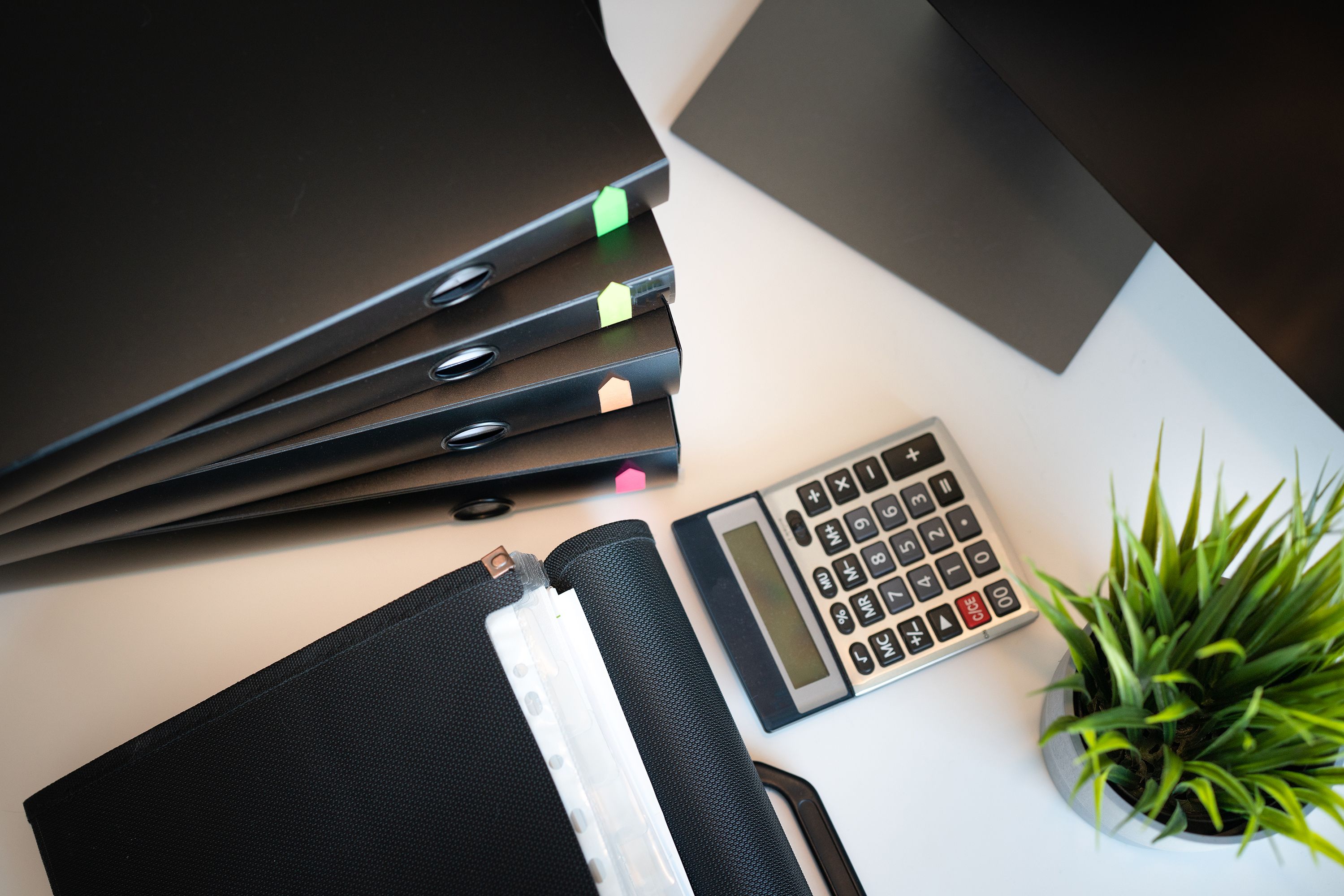 Stack of black binders with colored tabs, calculator, and small green plant on a white desk.
