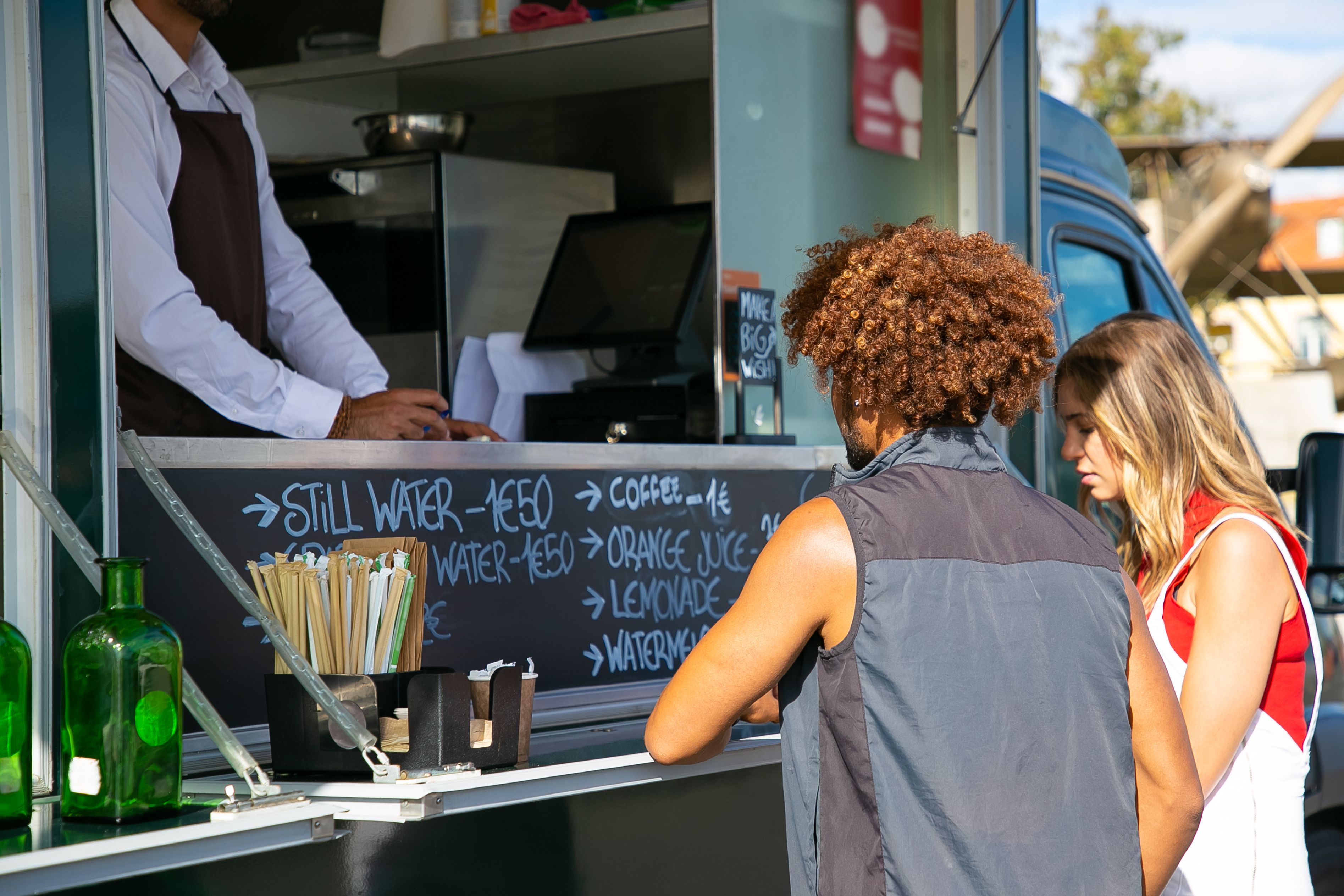 Two customers ordering drinks at an outdoor food truck with a handwritten menu and drink straws visible.