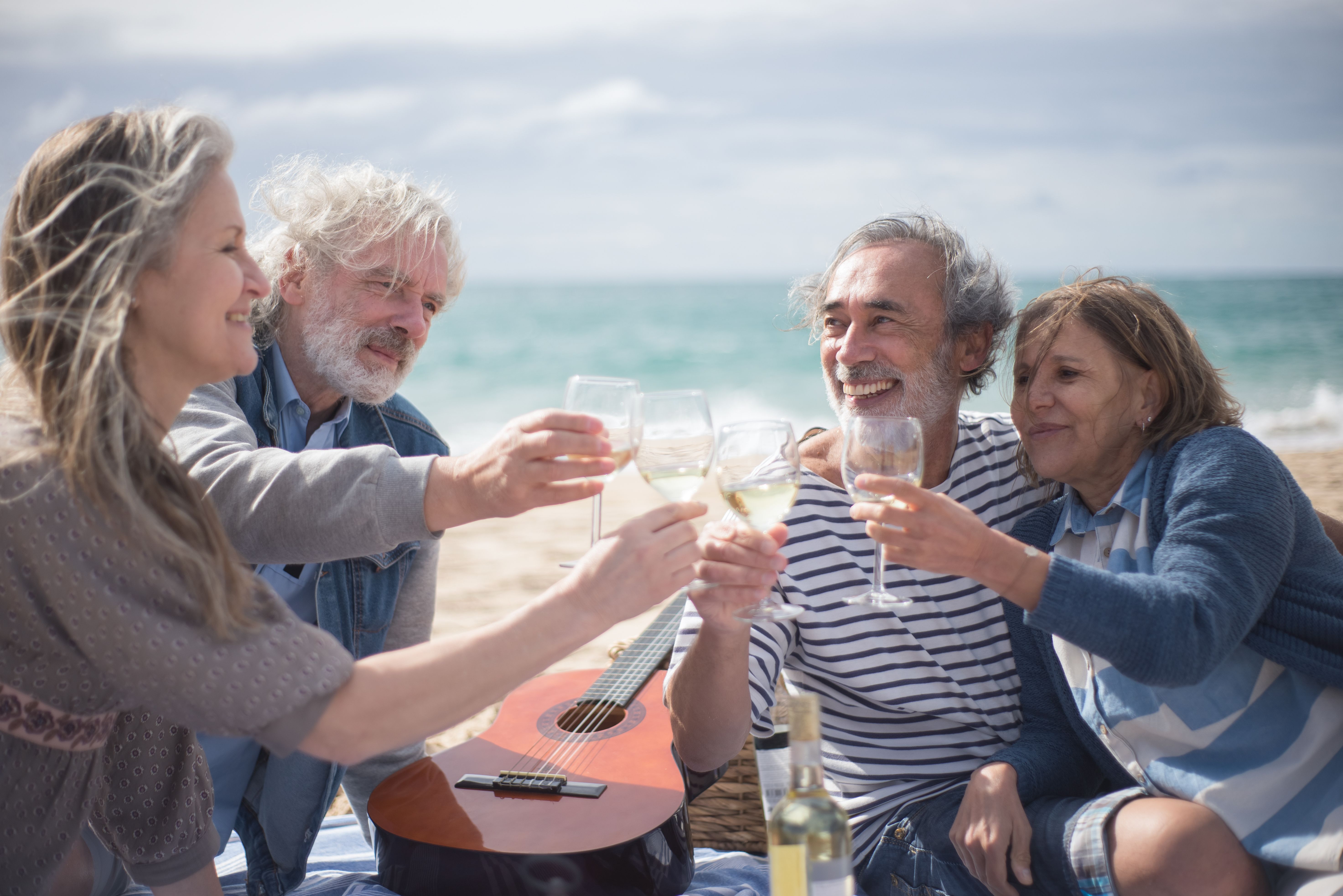 Four older adults clinking wine glasses and smiling on a beach with a guitar nearby.