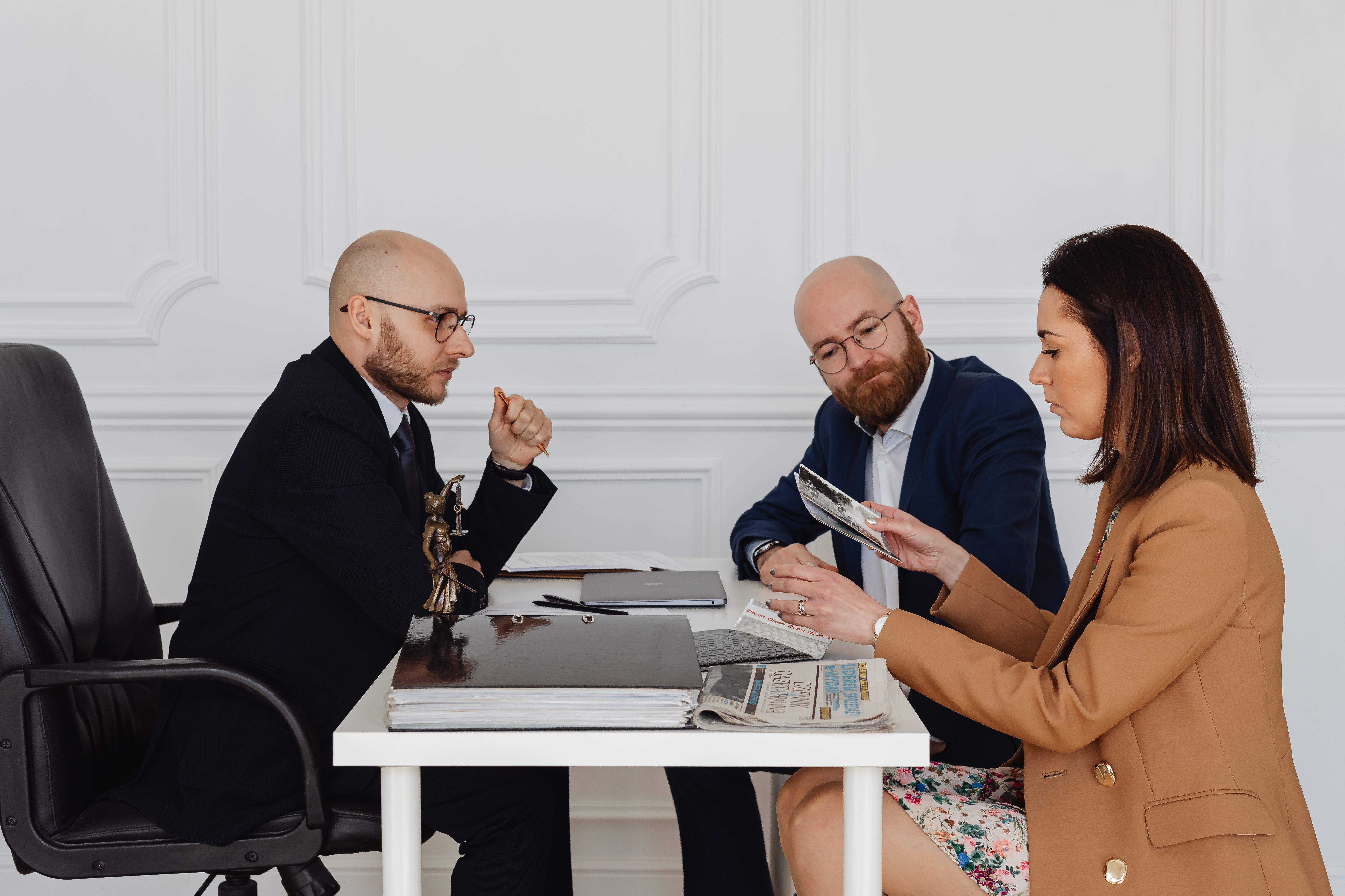 Three professionals at a table discussing documents and photos in a white office setting.