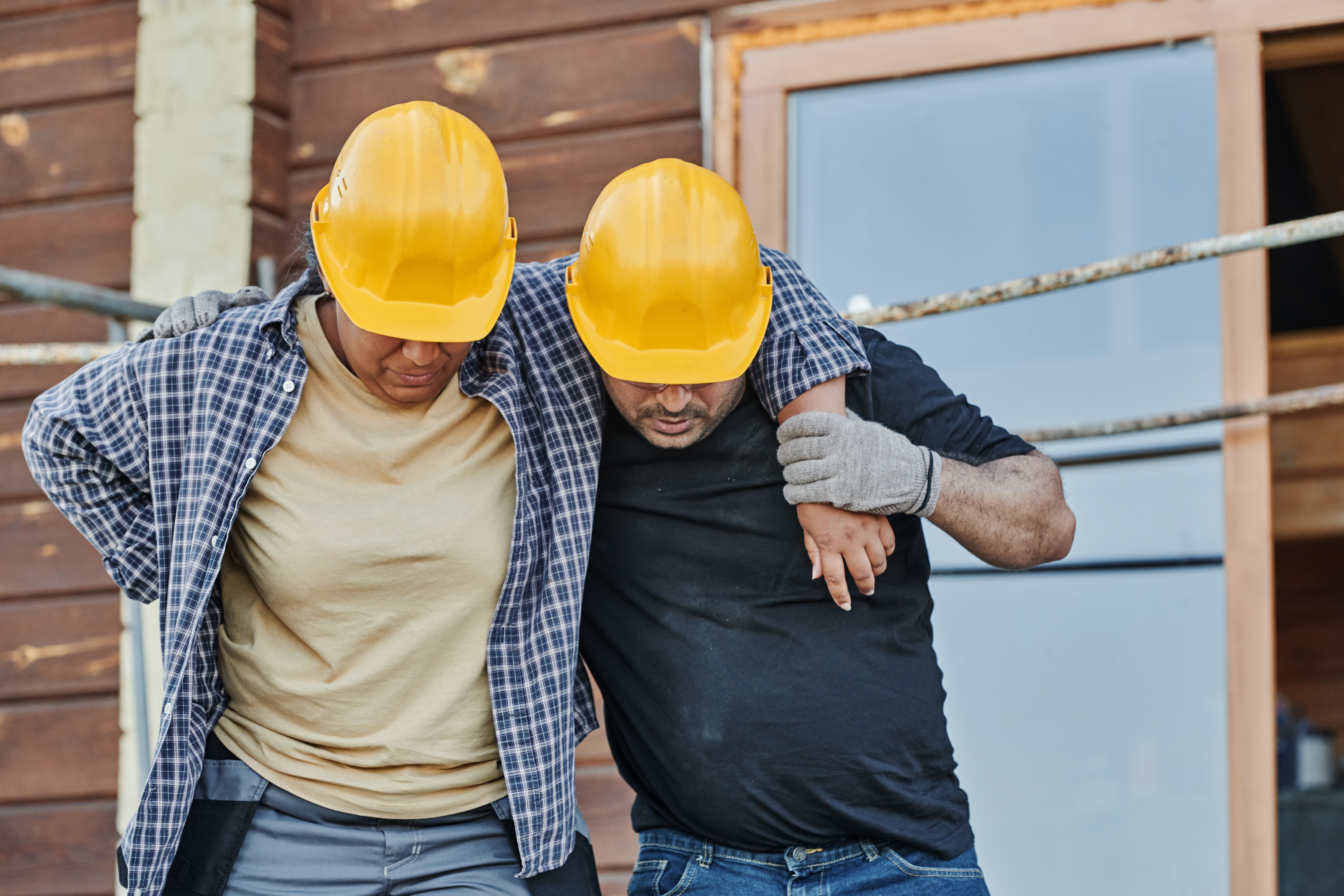 Two construction workers in yellow helmets support each other while walking at a building site.