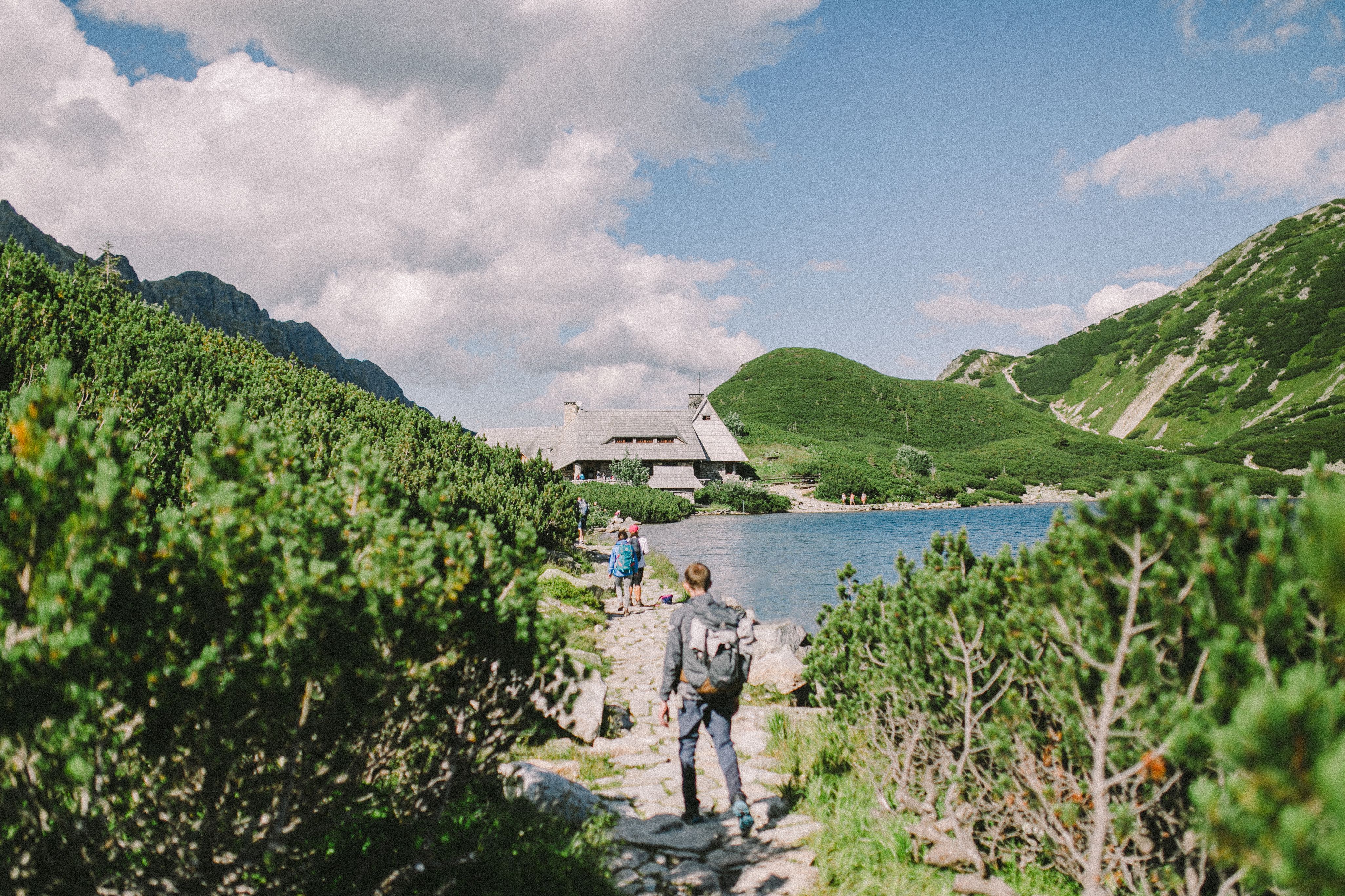 Hikers walking on a stone path beside a lake with mountains and a cabin in the background.