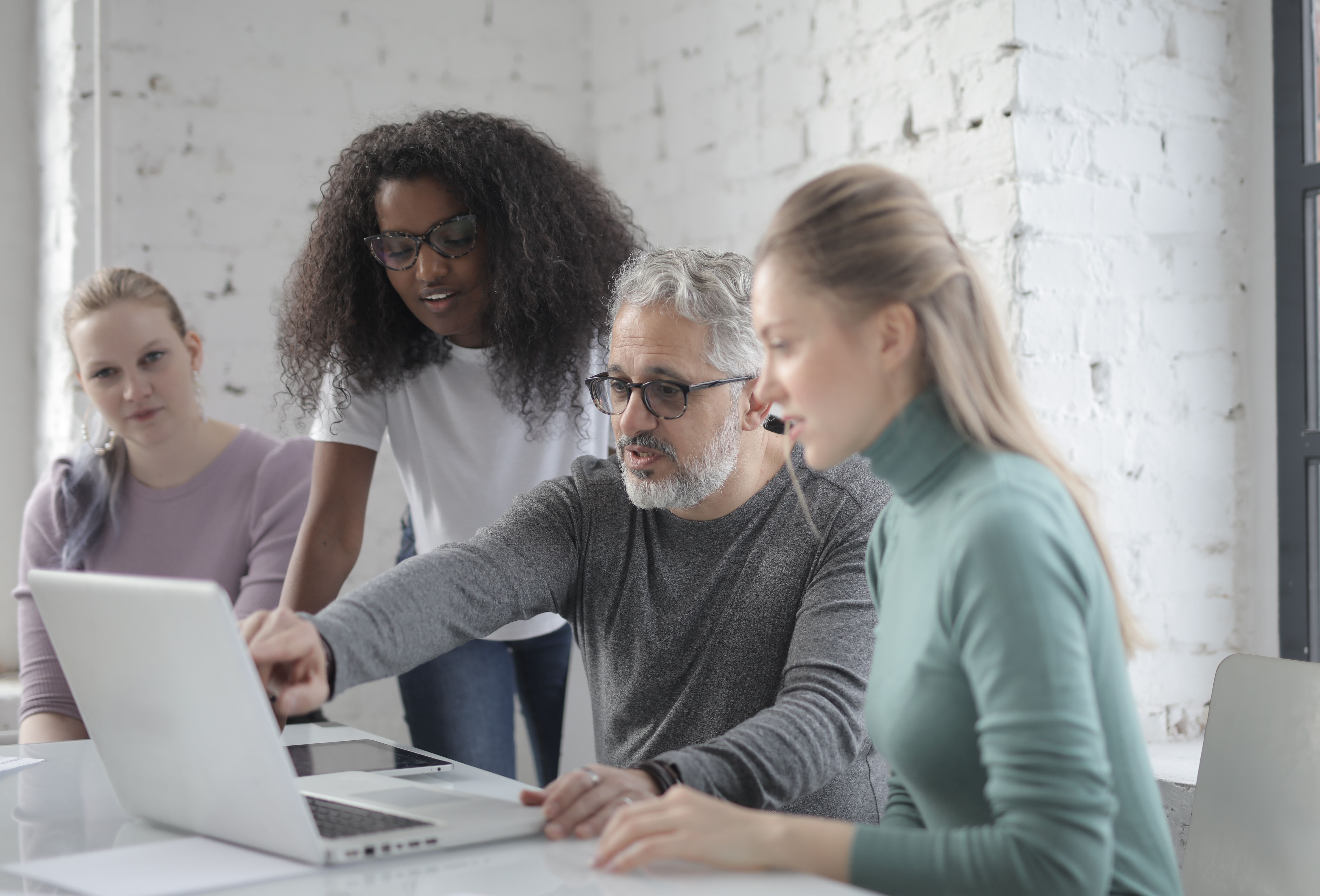 Four people collaborating and discussing something on a laptop in a modern office with white brick walls.