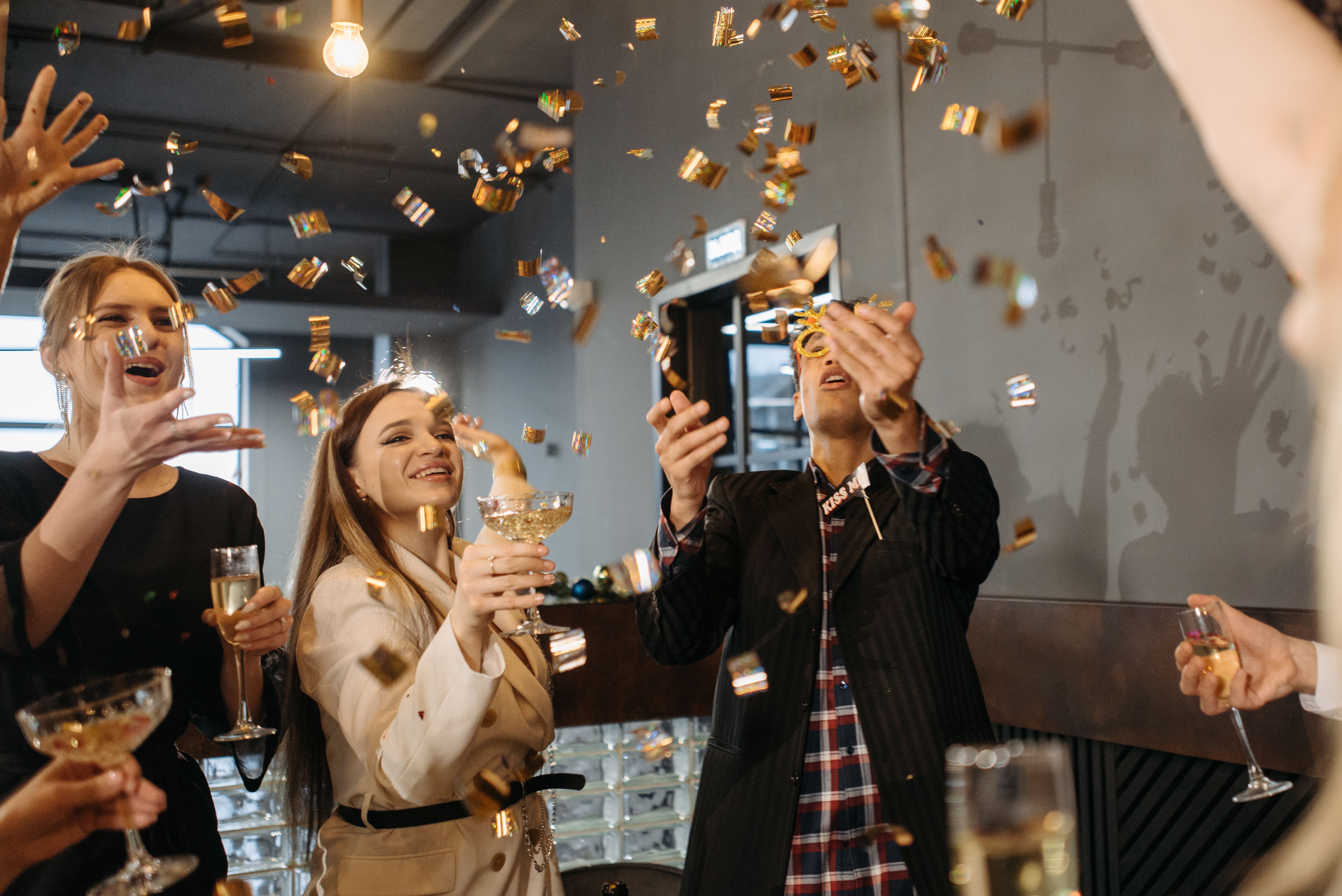 Group of young adults celebrating indoors with champagne and gold confetti falling around them.