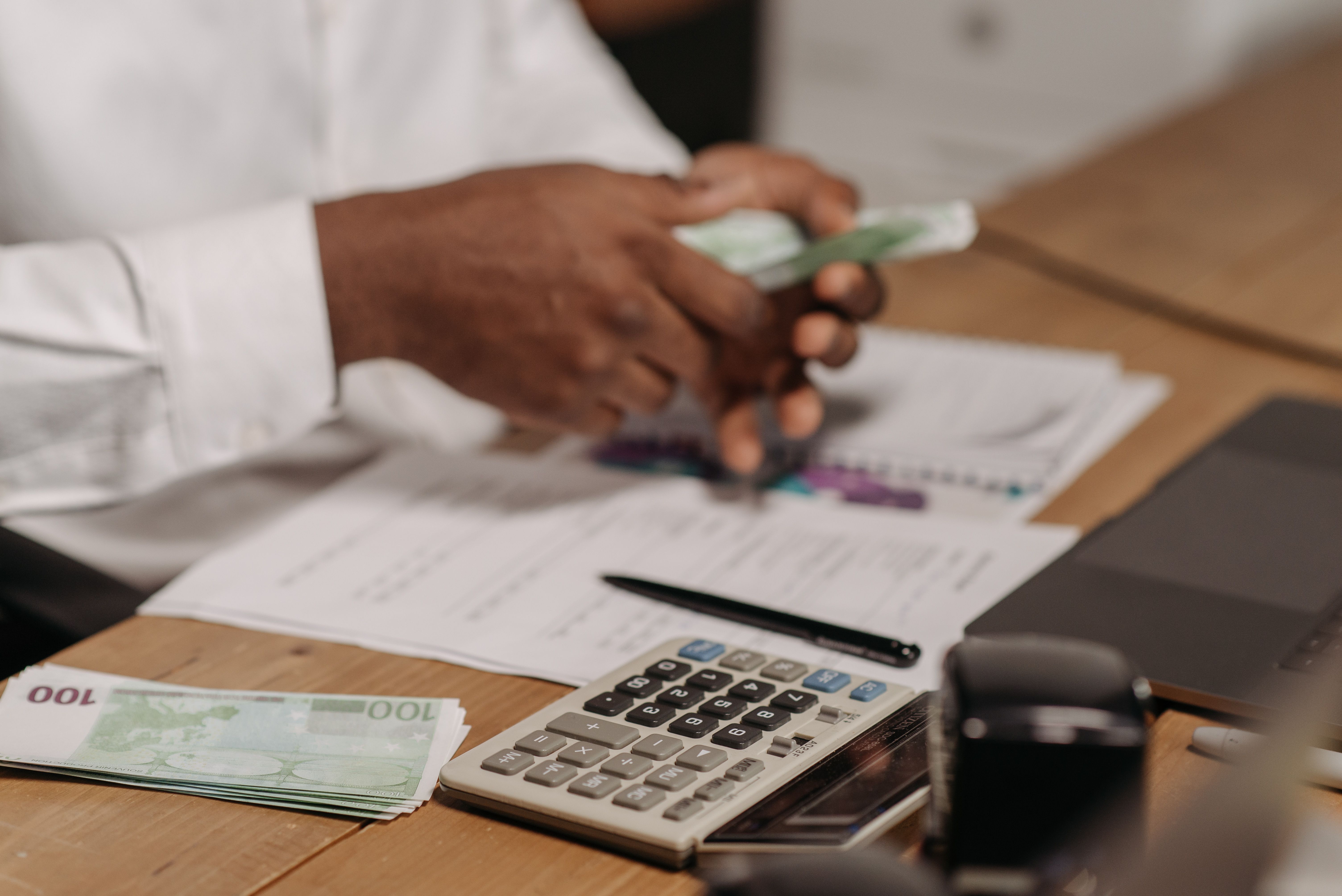 Person counting 100 euro bills at a desk with calculator, pen, documents, and laptop.