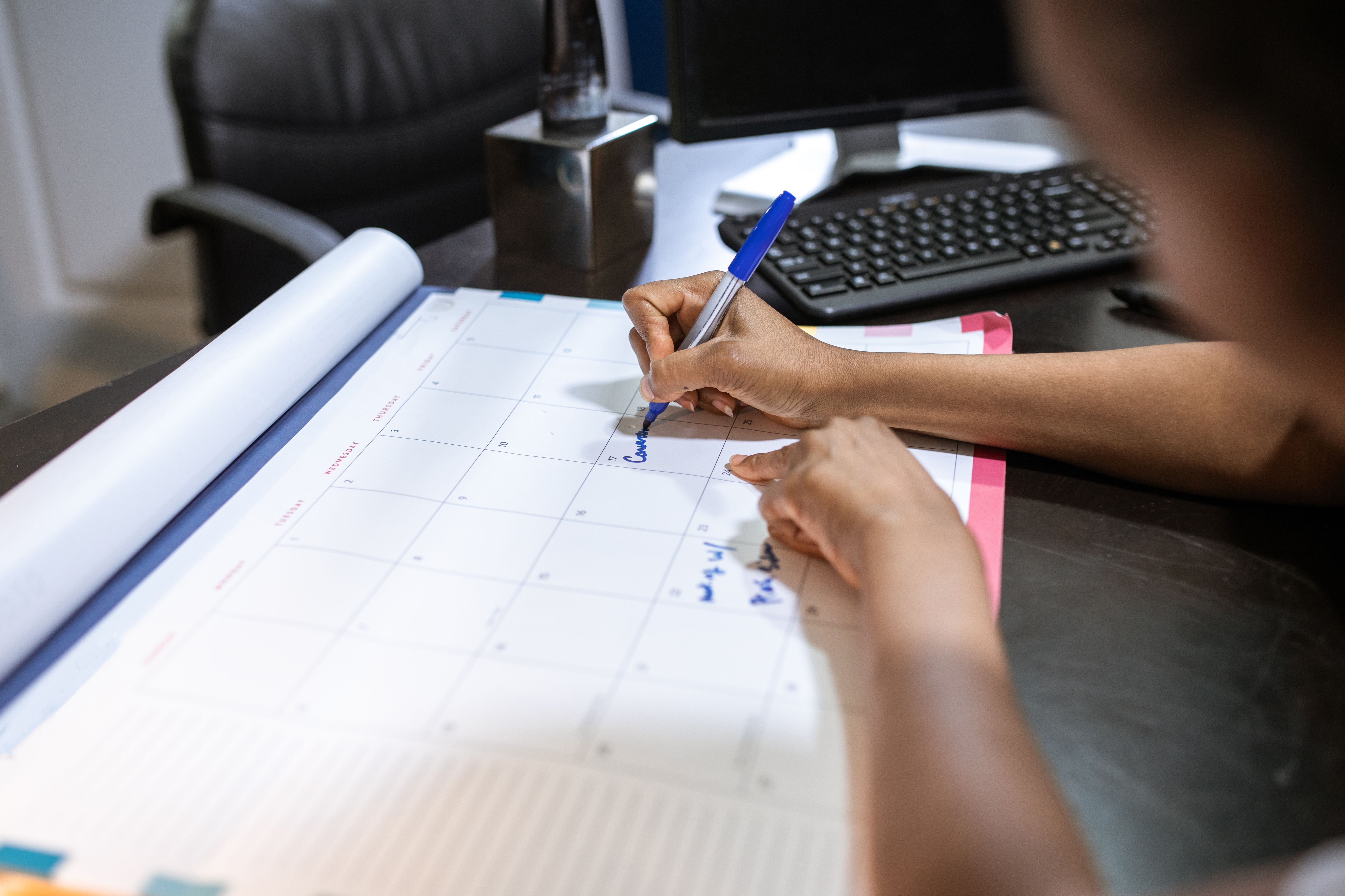 Person writing on a large desk calendar with a blue pen near a computer keyboard.