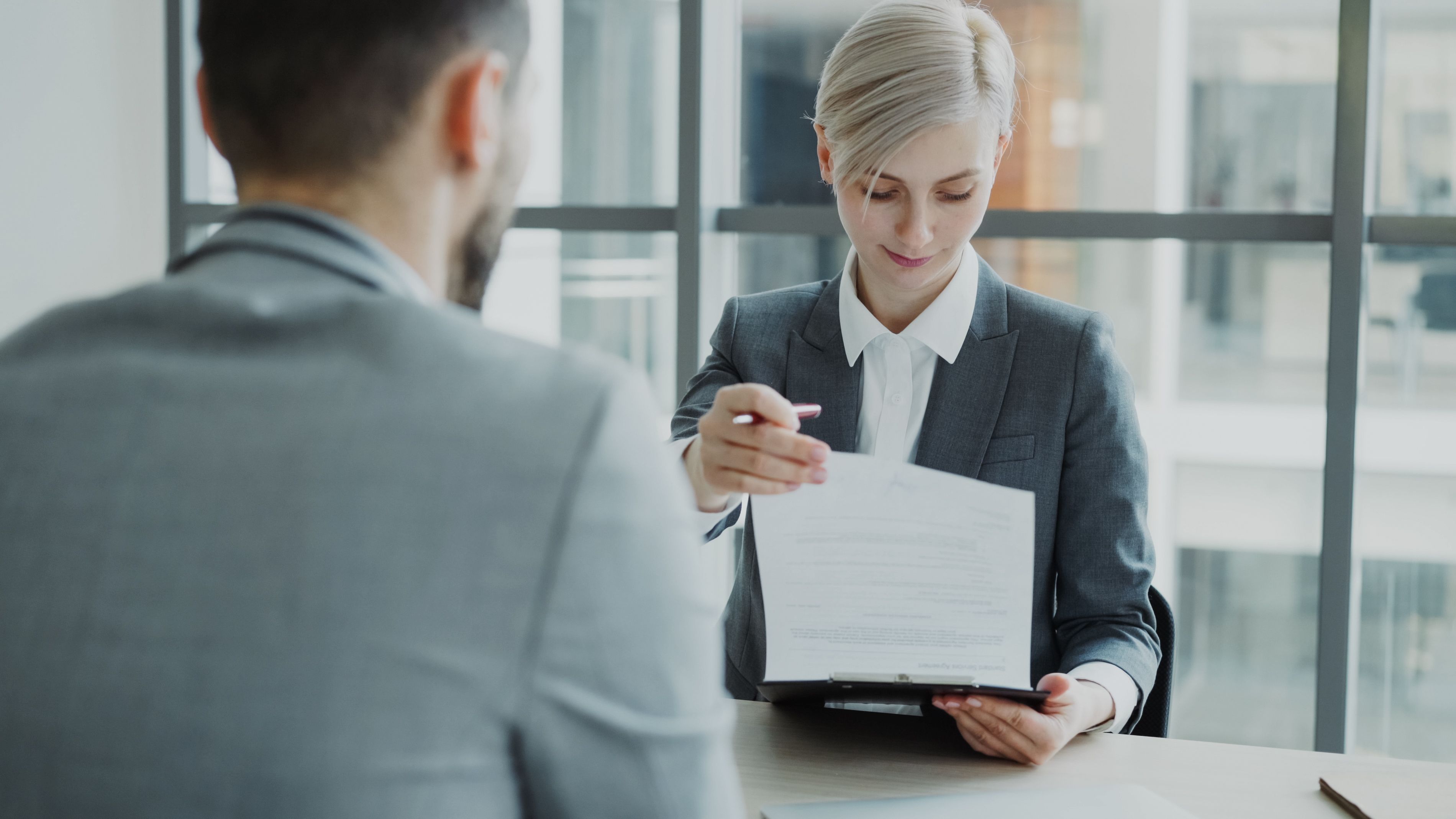Businesswoman in suit holding clipboard with document, showing it to man in office.