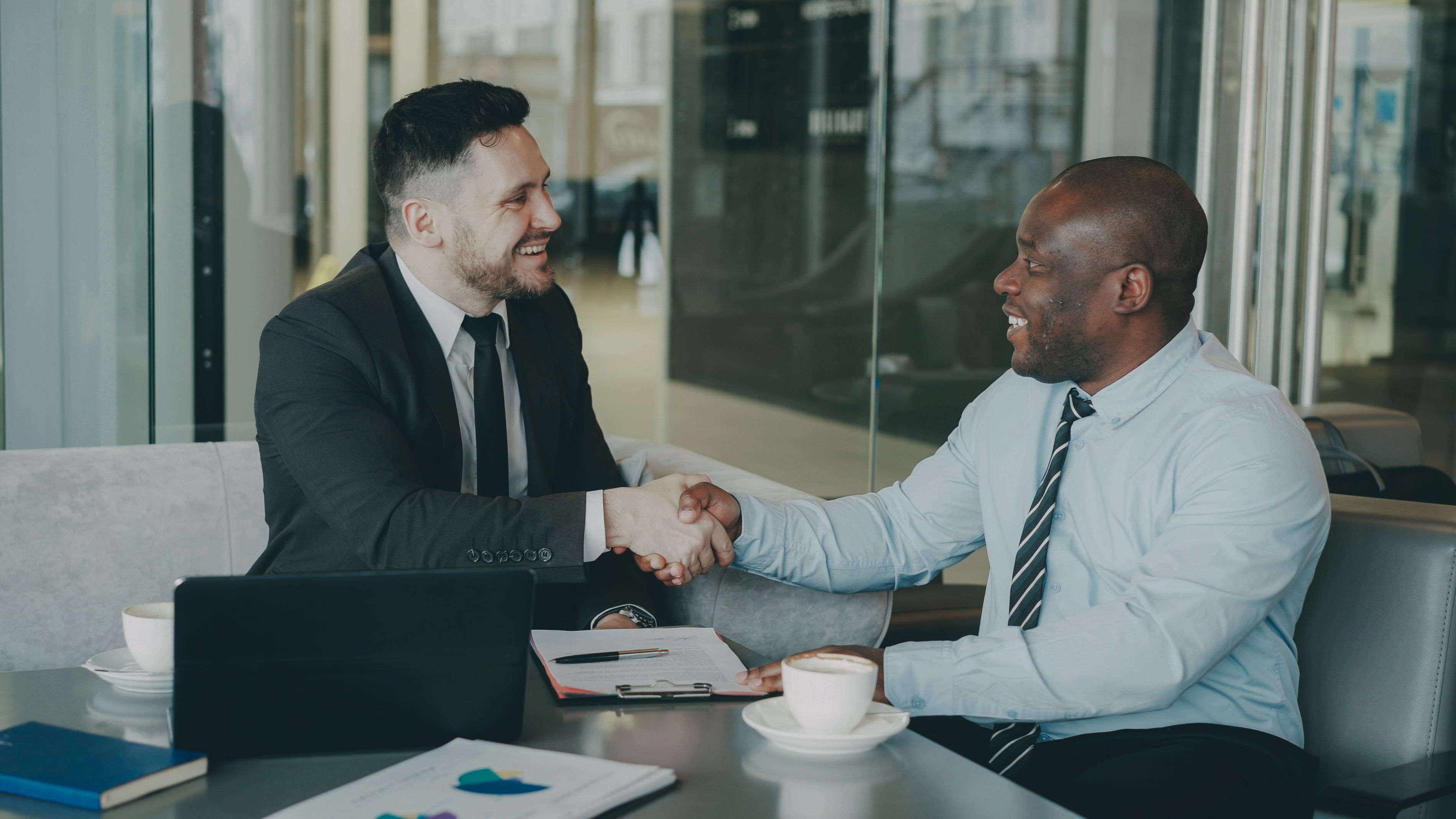 Two businessmen smiling and shaking hands across a table with coffee cups and documents.