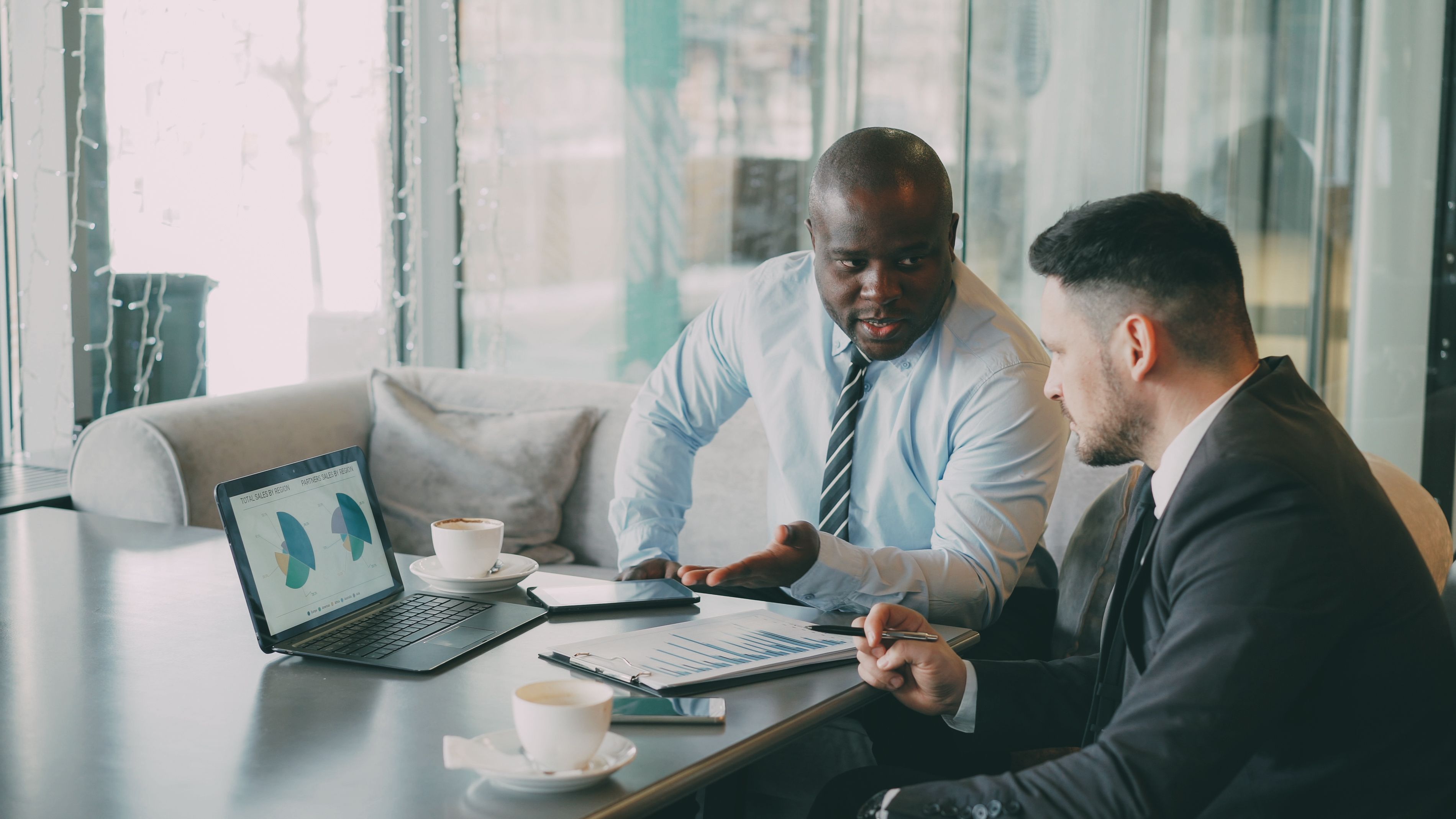 Two businessmen in suits discussing charts and graphs on a laptop and clipboard at a cafe table.