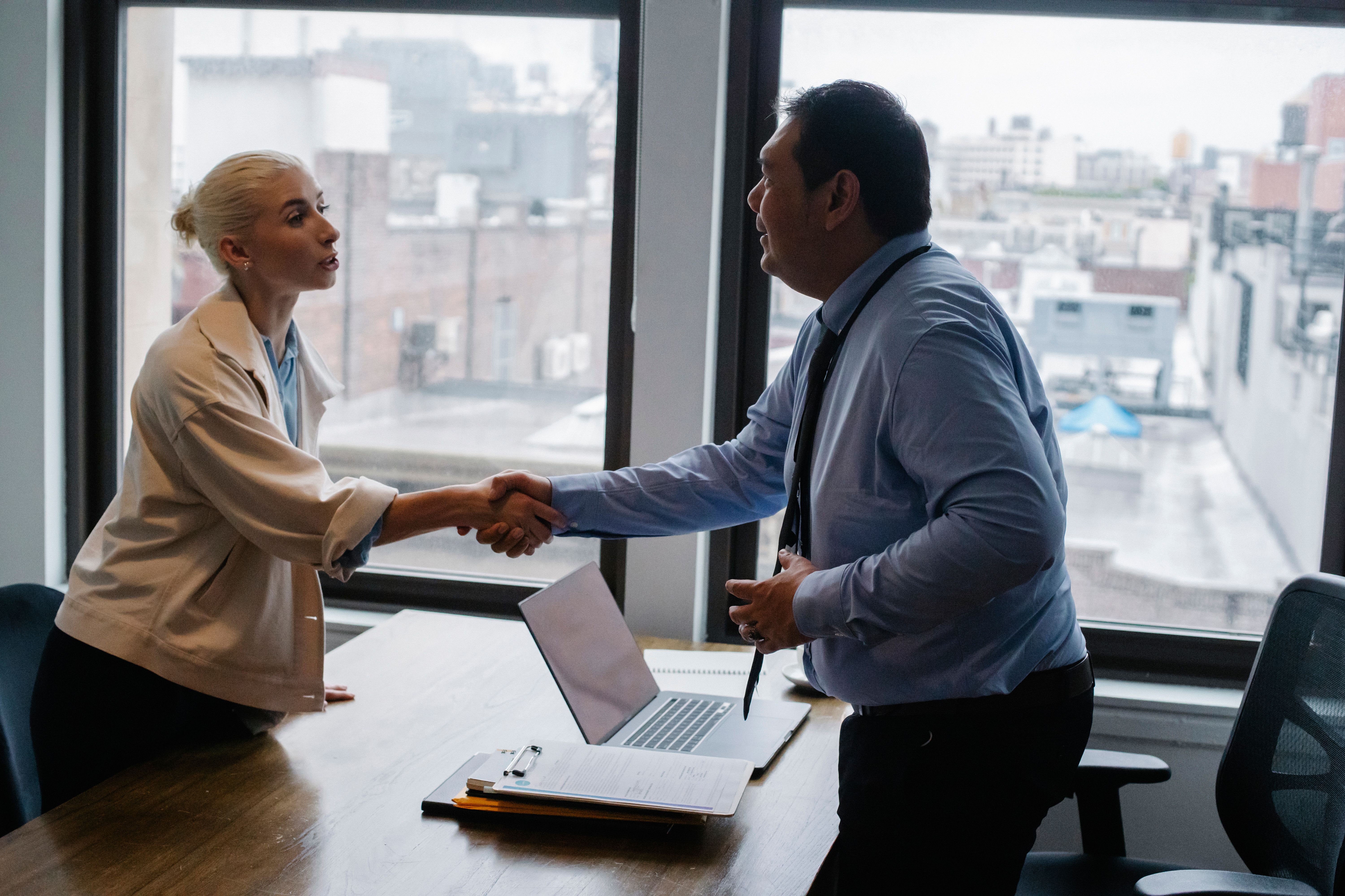 Two business professionals shaking hands across a desk with a laptop and documents in an office.