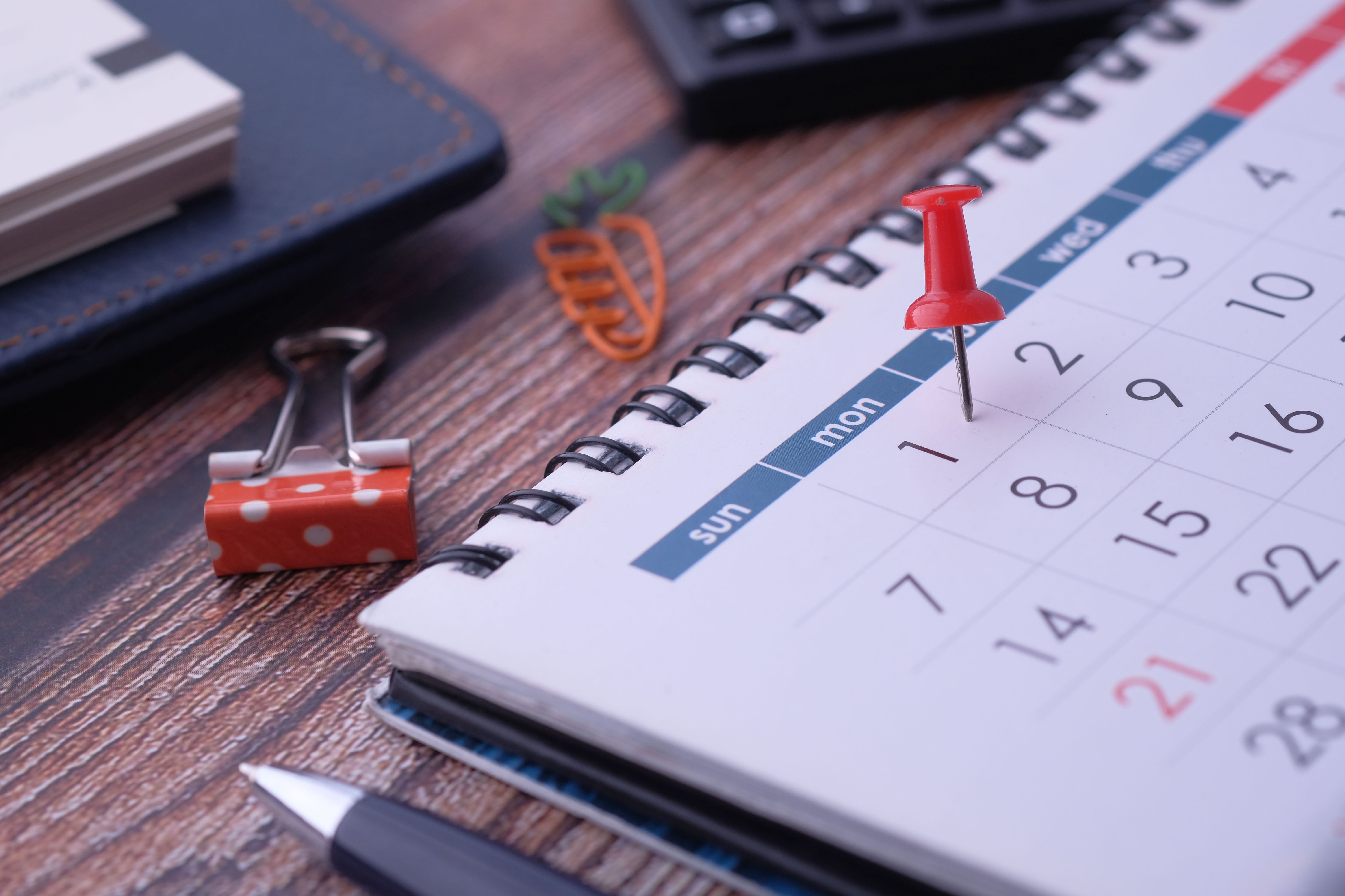 Red pushpin marking Monday the 1st on a spiral-bound calendar on a wooden desk.