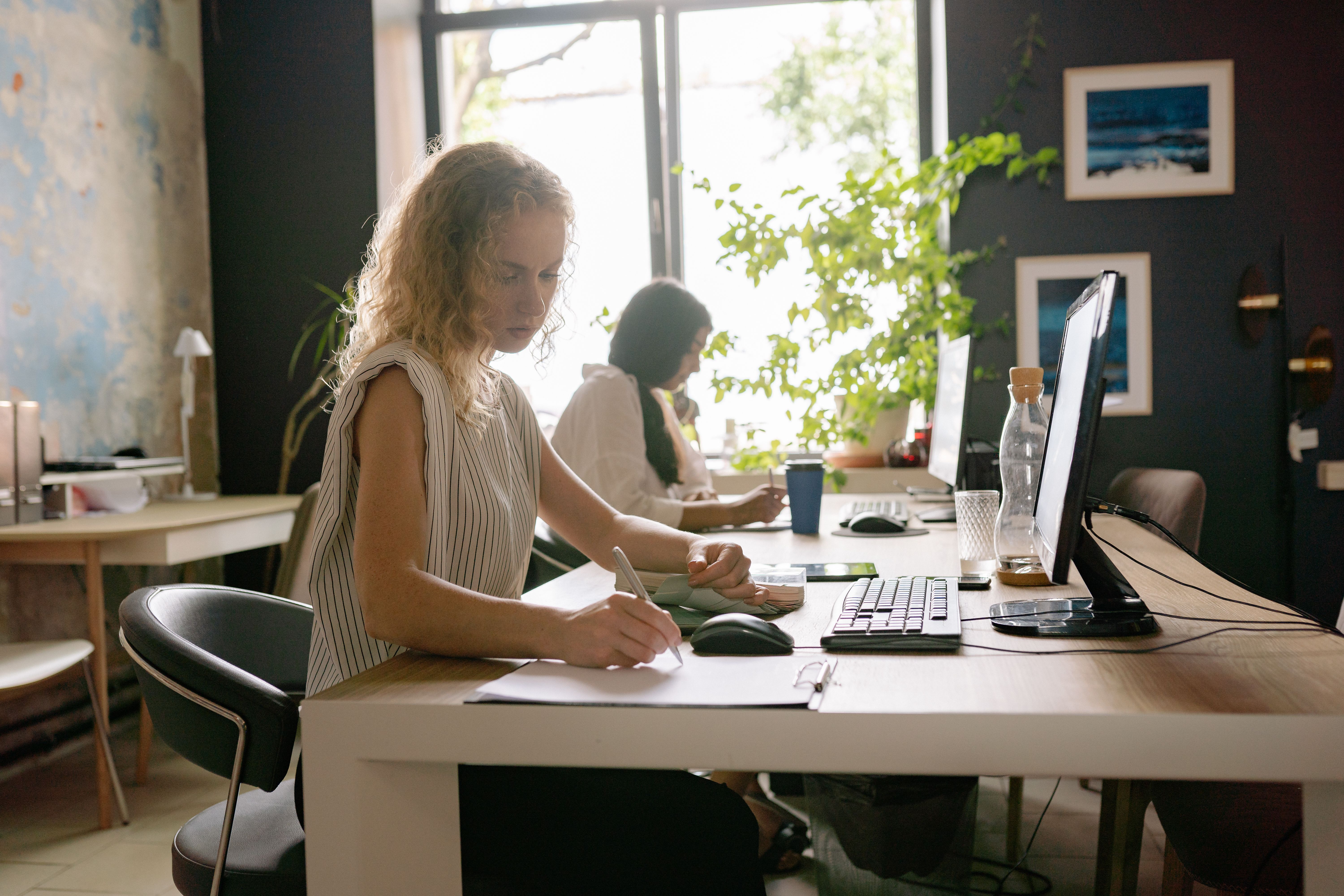 Two women working at desks, one writing on paper, the other looking at a device, in a bright office.