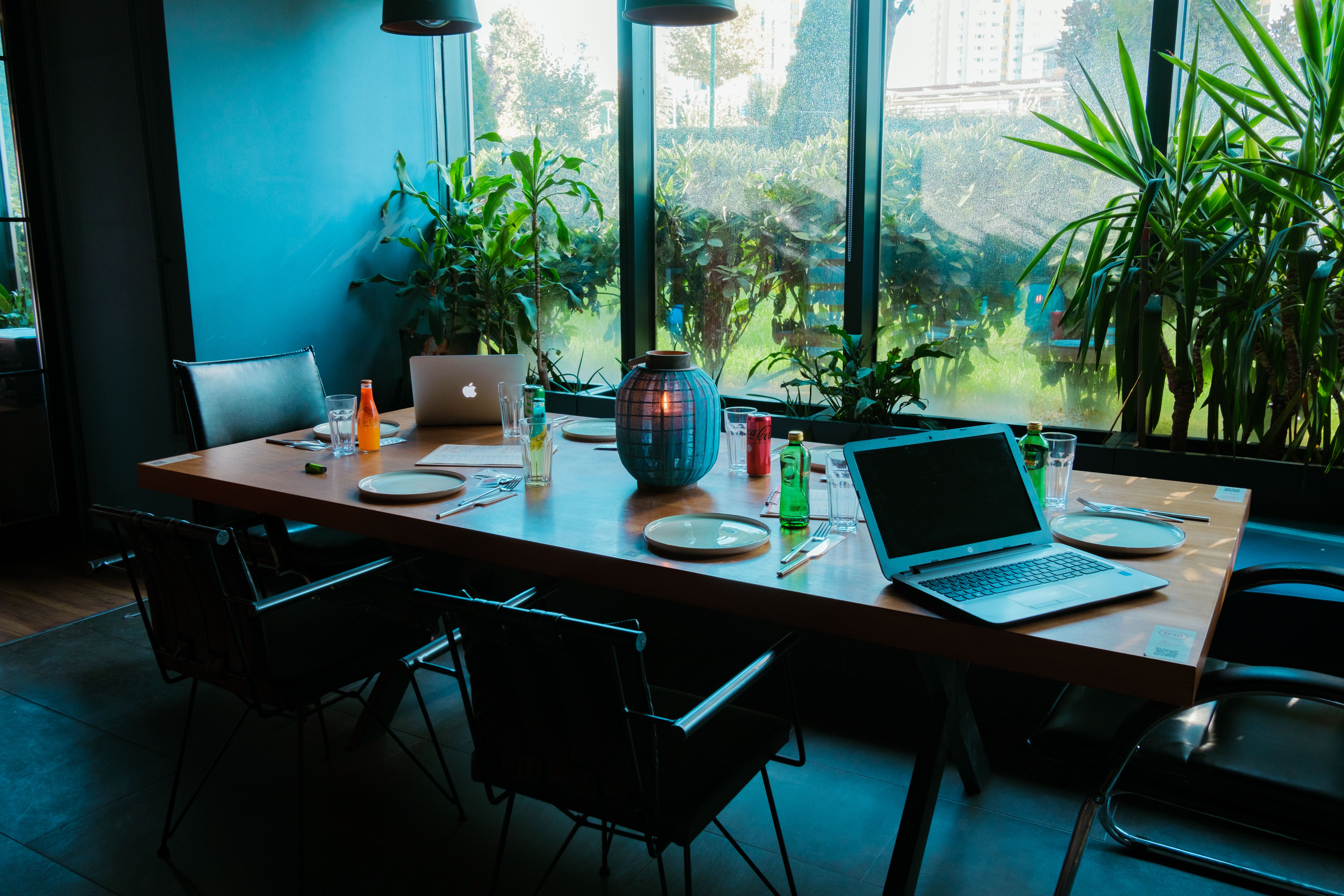 Wooden table with laptops, plates, drinks, and plants by a large window in a meeting room.